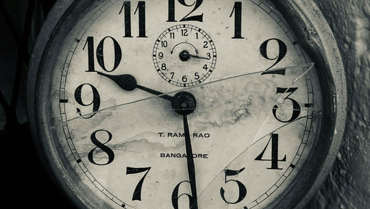 "Hilfe, ich habe keine Zeit!" - 5 Expertinnen-Tipps gegen Zeitmangel Alte, runde Wanduhr mit großen schwarzen Zahlen auf weißem Zifferblatt, das durch Risse und Flecken gezeichnet ist. Die Uhr zeigt die Zeit 10:29, mit einem kleineren Zifferblatt oben für Sekunden. Auf dem Zifferblatt steht "T. RAM RAO" und "BANGALORE".