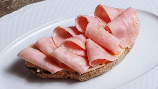 Jeden Tag Wurst essen: Das sind die Folgen für deinen Körper Offenes Sandwich mit mehreren dünn aufgerollten Scheiben gekochtem Schinken auf einer Scheibe Vollkornbrot, serviert auf einem weißen Teller.