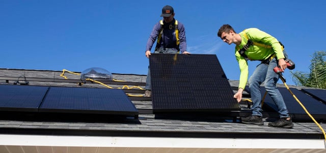 Zwei Männer installieren Solarpaneele auf einem Hausdach. Beide tragen Sicherheitsgurte, einer hält ein Solarpanel, der andere einen Akkuschrauber.