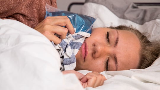 Warum werden Zahnschmerzen nachts oft schlimmer? Frau liegt im Bett und hält eine blaue Kältekompresse mit einem Tuch an ihre Wange, die Augen sind geschlossen.