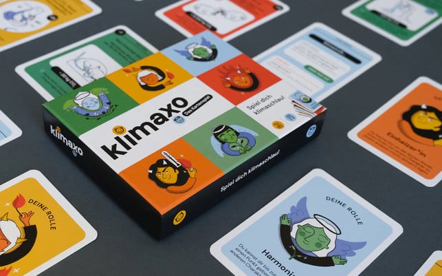 klimaxo Spielkartenspiel "klimaxo" mit farbigen Karten, die verschiedene Rollen und Aktionen zum Thema Klimaschutz darstellen. Die Verpackung zeigt bunte Illustrationen von Figuren mit Symbolen wie Flammen, Thermometer und Engelsflügeln.