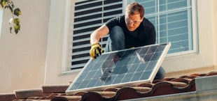 Ein weißer Mann mit Schutzbrille und Arbeitshandschuhen installiert ein Solarpanel auf einem Dach mit roten Ziegeln .