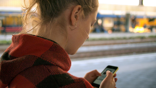 Eine Frau bedient ihr Smartphone. Sie steht an einem Bahnsteig. Eine Frau bedient ihr Smartphone. Sie steht an einem Bahnsteig.