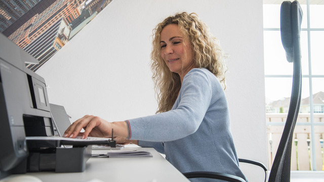 Klimaanlage von der Steuer absetzen: Unter diesen Bedingungen klappt das Frau mit lockigem, blondem Haar sitzt an einem Schreibtisch und legt ein Blatt Papier in einen Drucker. Sie trägt einen hellblauen Pullover und sitzt auf einem schwarzen Bürostuhl vor einer weißen Wand mit einem großen Fenster im Hintergrund.