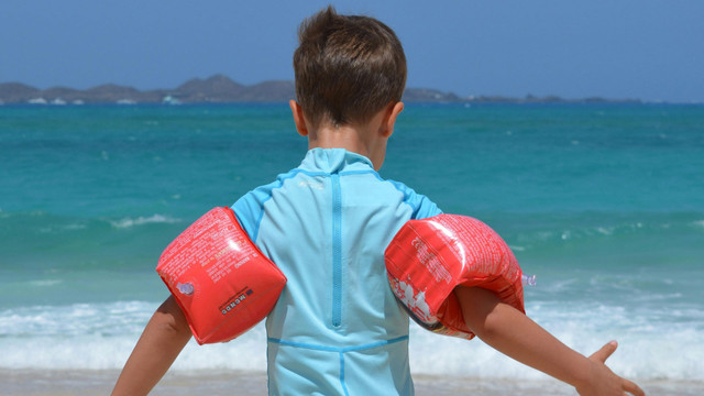 Gebrauchte UV-Kleidung kann weniger schützen – worauf Eltern achten müssen Ein Kind mit kurzen braunen Haaren trägt ein hellblaues Schwimmshirt und rote Schwimmflügel an beiden Armen, steht am Strand und blickt aufs Meer mit blauem Wasser und Bergen im Hintergrund.