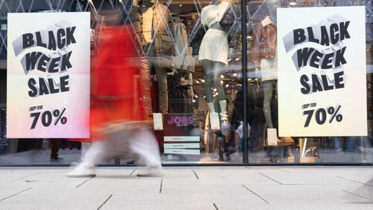 "Dauersuche nach einem Schnäppchen-Kick": Was längere Rabattschlachten in uns bewirken Schaufenster eines Bekleidungsgeschäfts mit zwei großen Plakaten, die "Black Week Sale up to 70%" ankündigen. Im Hintergrund sind mehrere Schaufensterpuppen mit Winterkleidung zu sehen. Vor dem Schaufenster läuft eine Person in rotem Mantel und weißer Hose, die durch Bewegungsunschärfe verschwommen ist.