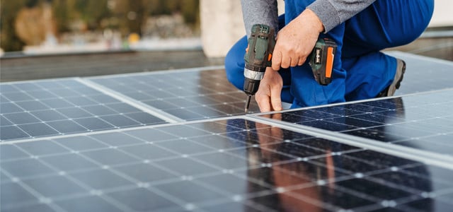 Eine Person in blauer Arbeitskleidung und grauem Pullover montiert Solarpaneele auf einem Dach und benutzt dabei einen Akkuschrauber.