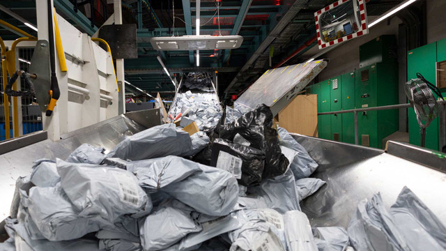 EU will 2026 Abgabe auf Billig-Importe einführen: wichtiger Schritt gegen Temu und Co. Internationales Postzentrum (IPZ) am Frankfurter Flughafen: Förderband mit zahlreichen verpackten Paketen in grauen und schwarzen Kunststoffhüllen in einer Lager- oder Sortieranlage, im Hintergrund grüne Schaltschränke und eine Überwachungsspiegel an der Wand.