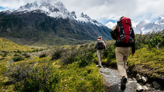 Welche Outdoor-Kleidung brauche ich wirklich? "Für den Anfang reichen wenige Teile" Zwei Wanderer mit Rucksäcken auf einem schmalen Pfad in einer bergigen Landschaft, im Hintergrund schneebedeckte Gipfel unter bewölktem Himmel.