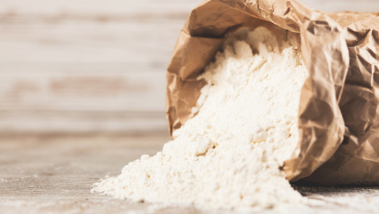mehl-vollkornmehl-cb-Georgii-Dolgykh-260323 Eine geöffnete braune Papiertüte, aus der weißes Mehl auf eine helle Holzoberfläche rieselt.